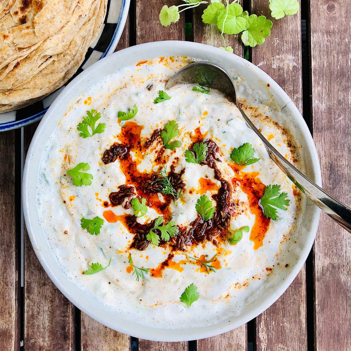 A bowl of cucumber raita garnished with parsley on a slatted wooden table