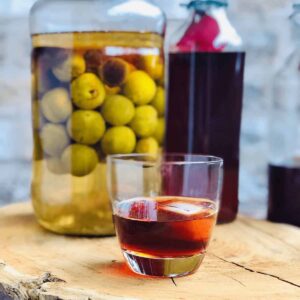 A glass of umeshu plum wine with a large jar of plums infusing, and a bottle of plum wine in the background