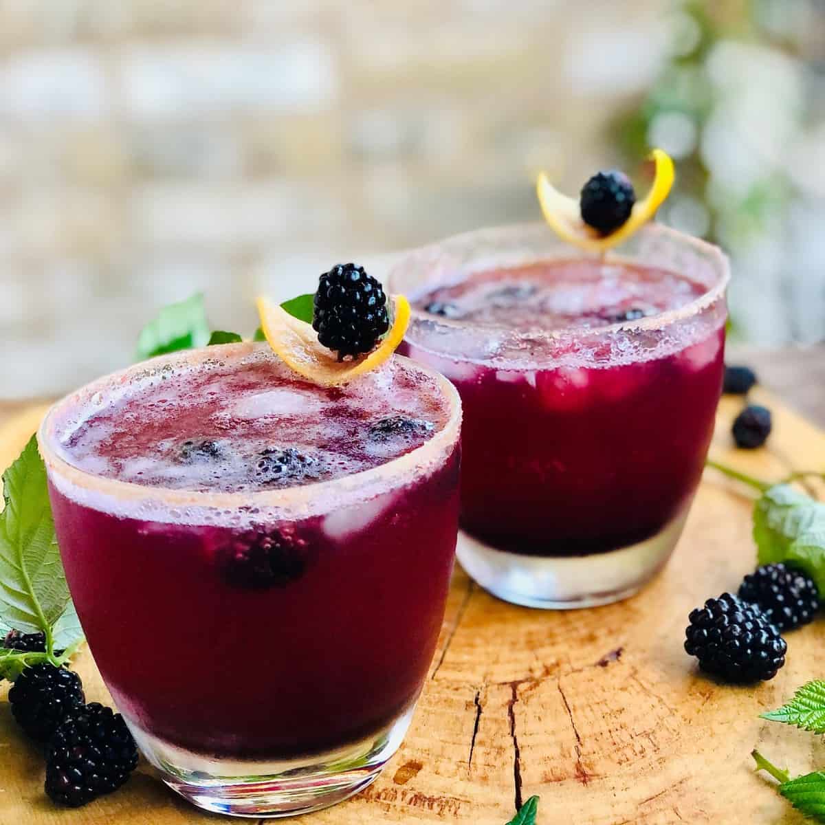 Two glasses of Kala Khatta mocktail garnished with lemon slice and a blackberry.
