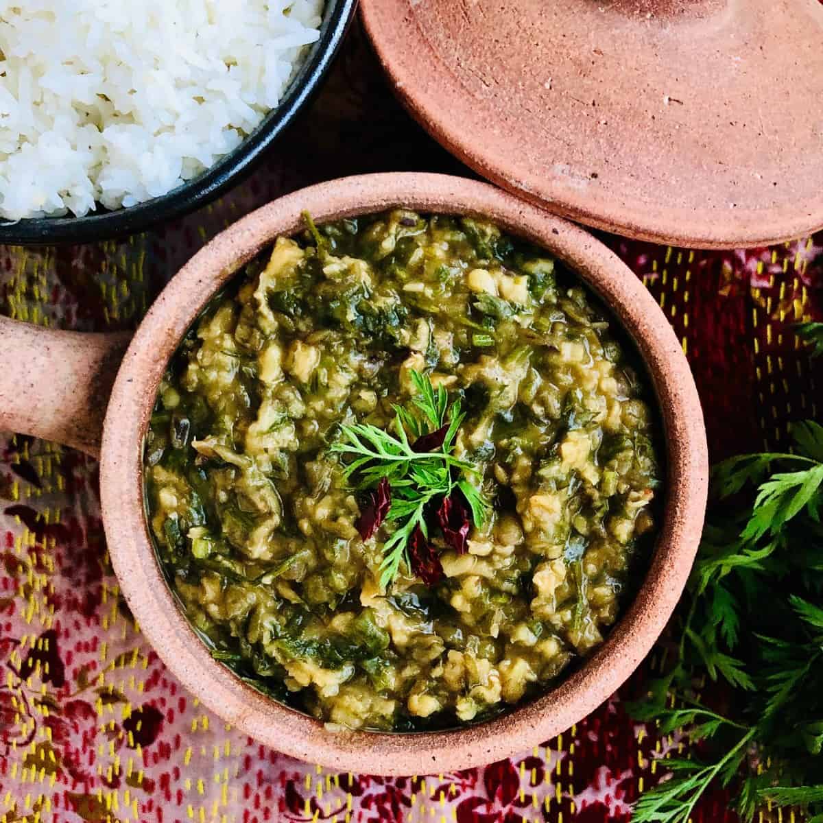 A clay pot containing saag daal garnished with carrot tops. 
