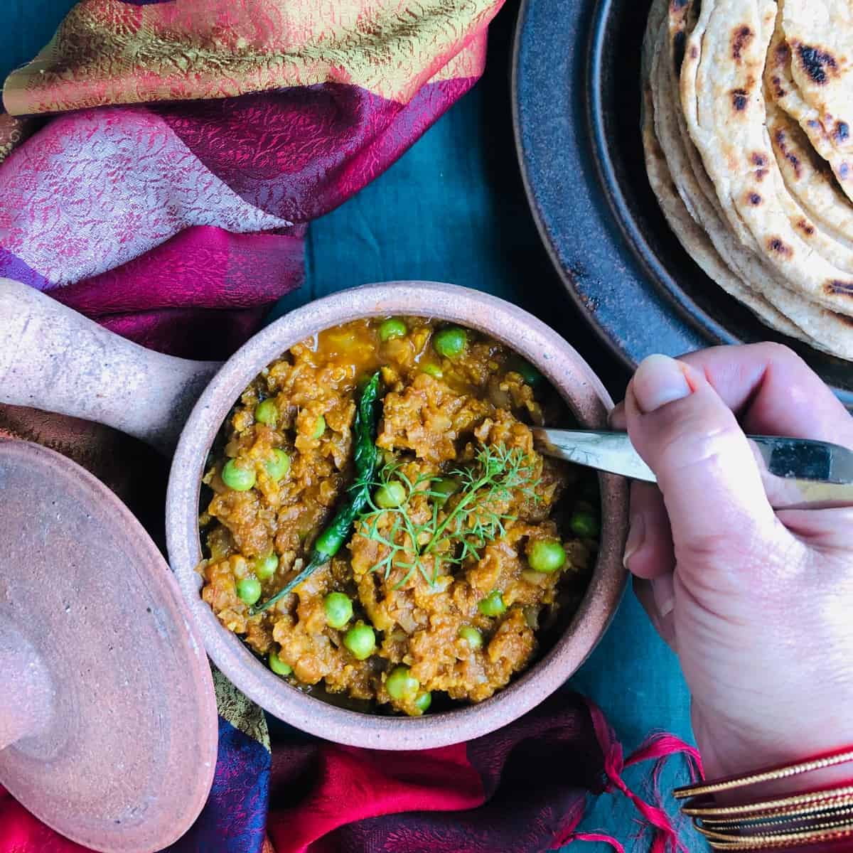 Clay pot containing vegan mince Keema Mattar. A hand holds a spoon in the keema mattar on the right hand side.