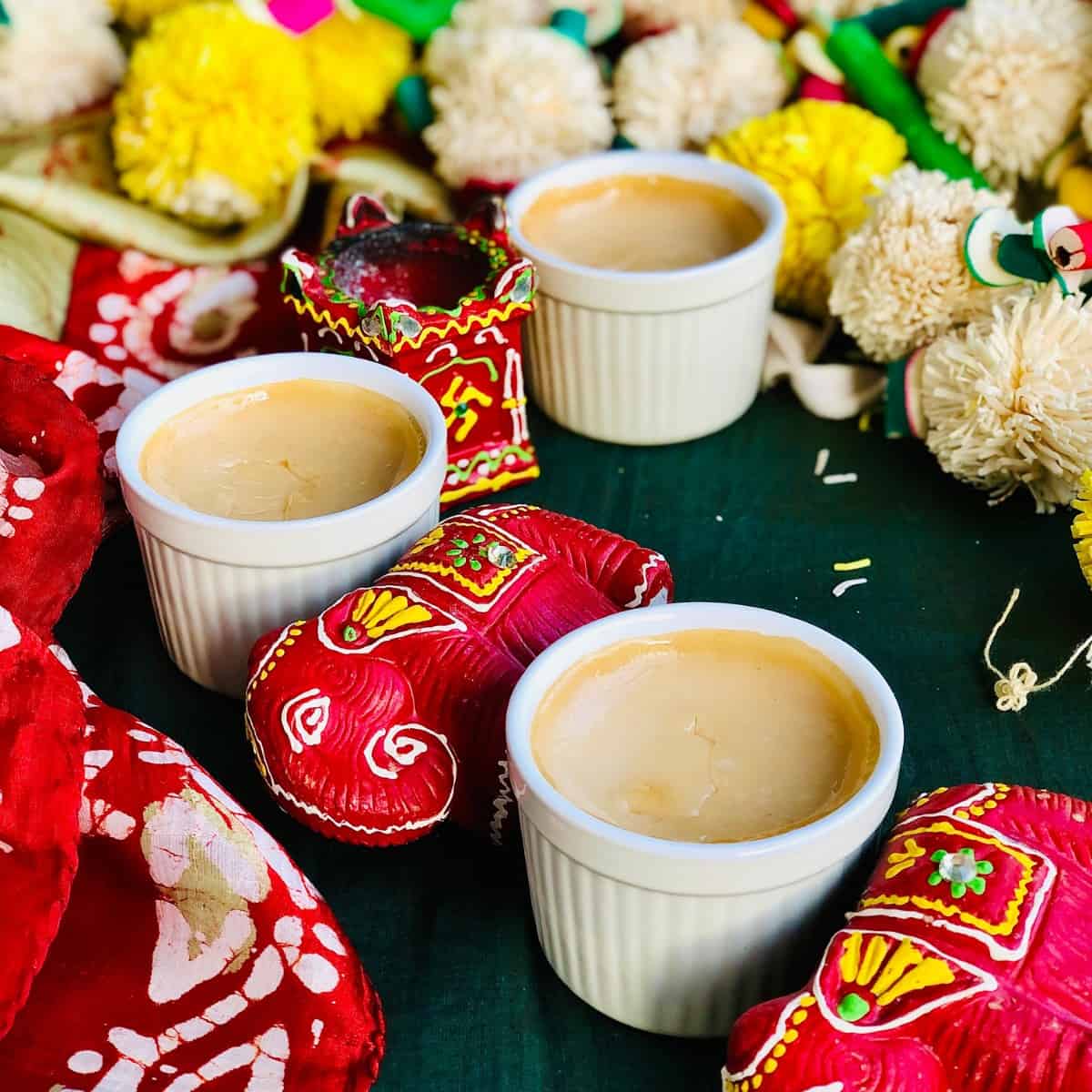 Three small pots of bhapa doi indian dessert sitting on a dark green cloth surrounded by diwali decorations.
