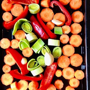 A baking tray containing sliced raw carrots, red pepper and leek