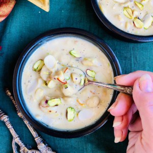 A dish of makhana beer garnished with slivers of pistachio. A hand holds a spoon full of makhana keer over the dish. 