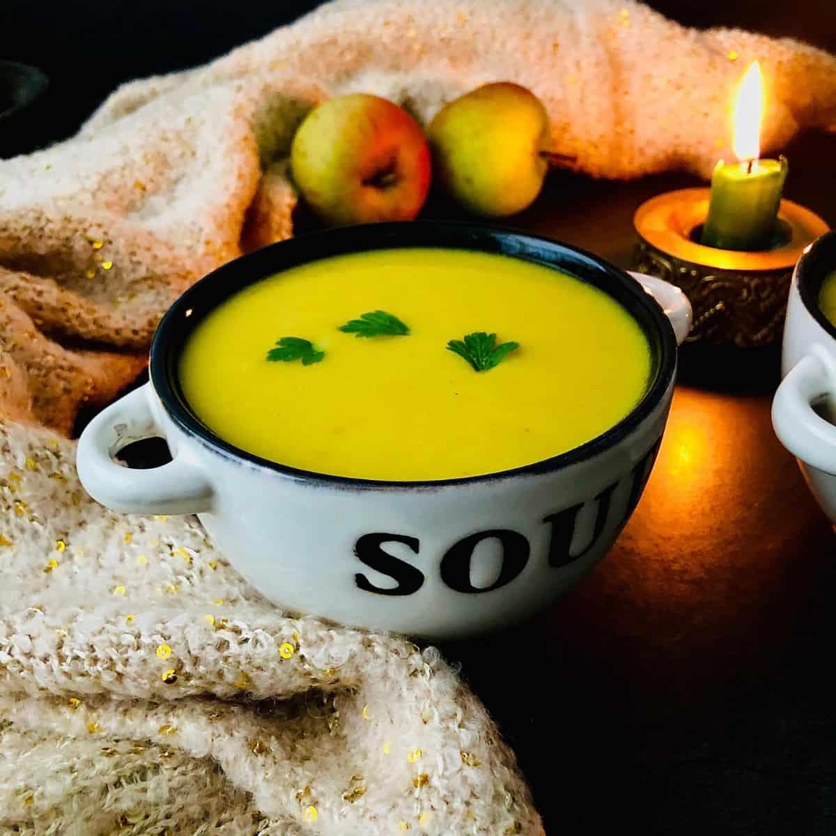 Bowl of Mulligatawny soup. The bowl sits nestled in a soft wool fabric. A lit candle and to whole apples sit in the background as decoration.