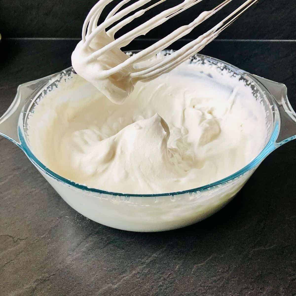 A bowl containing whipped vegan cream. A whisk is held over the bowl
