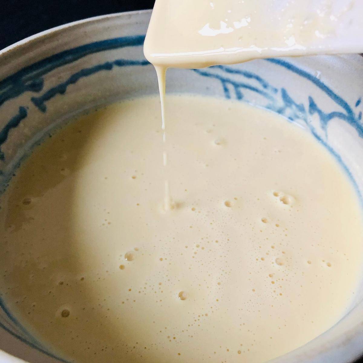 Gram flour paneer batter after water added. The batter flows off a spatula above the bowl to show consistency