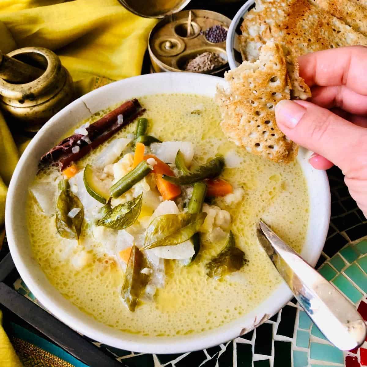 A bowl of ishtew vegetable stew. A hand hovers over the bowl holding a piece of dosa
