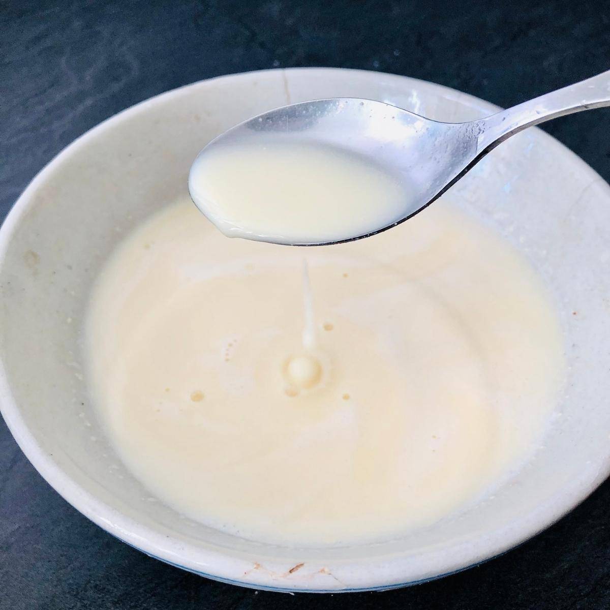 A bowl containing gram flour, yoghurt and water mix. A spoon hovers over the bowl with the mix dropping into the bowl