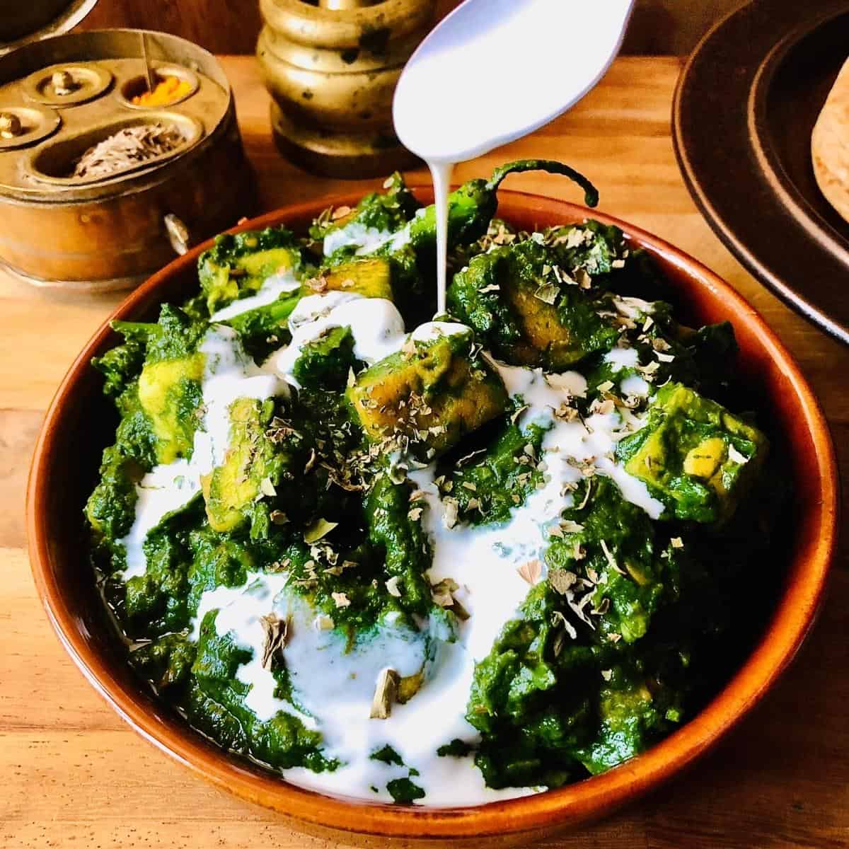 A dish of palak paneer with a spoon pouring yoghurt over the palak paneer
