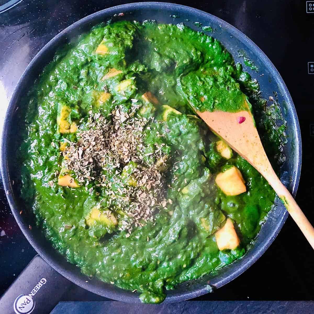 Fenugreek added to palak paneer mix in a frying pan. A wooden spoon rests in the pan
