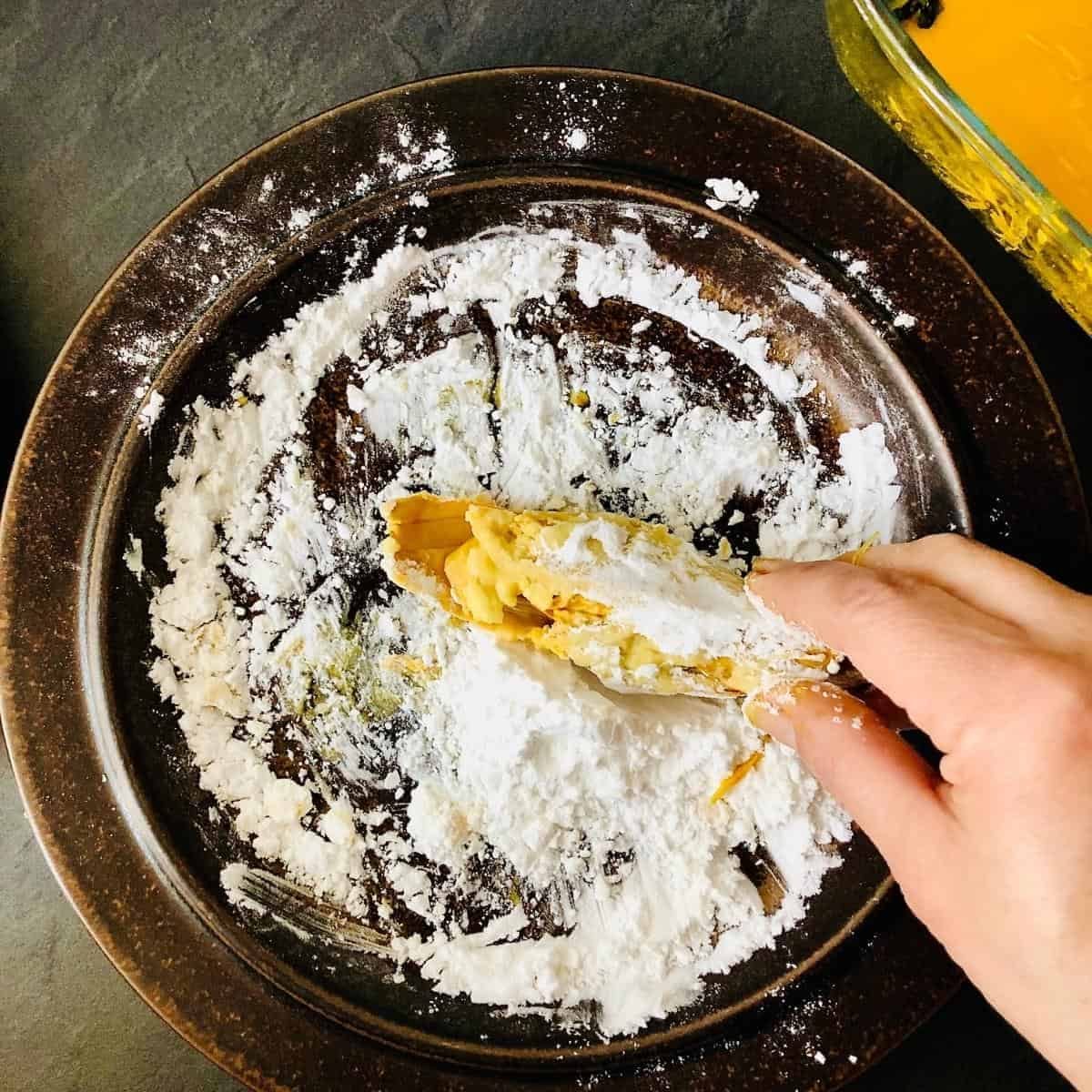 Brown plate containing potato starch. A hand holds a banana blossom in the potato starch, coating it.