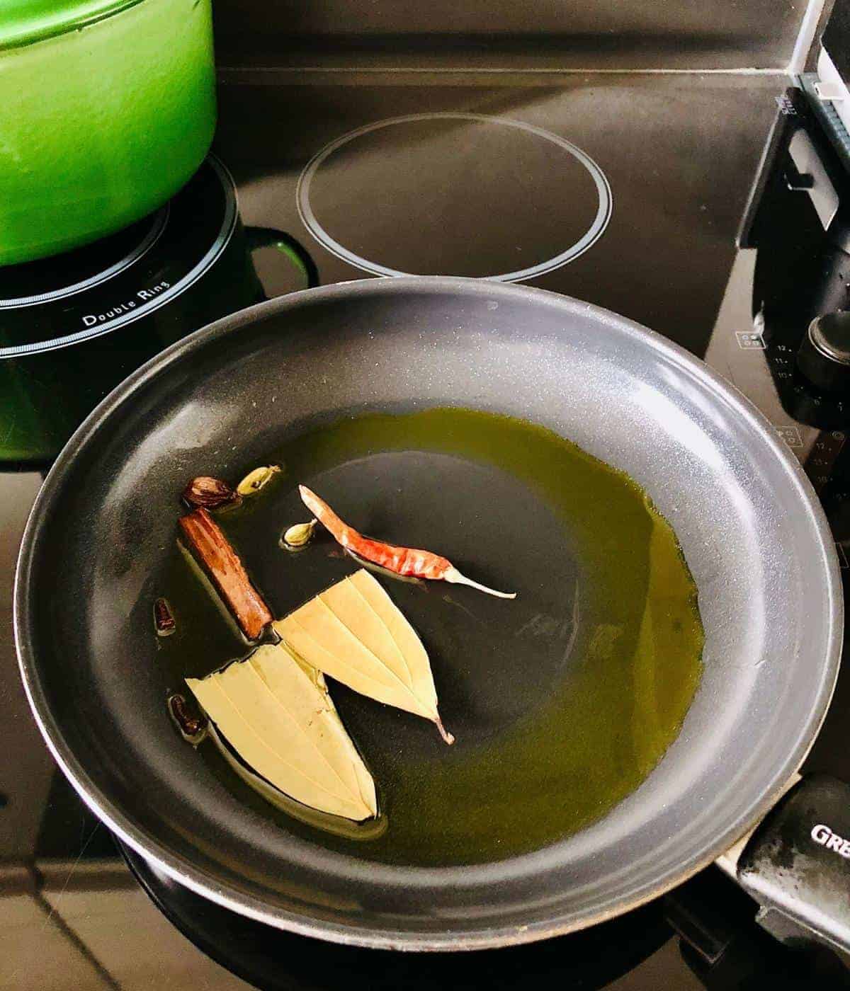 Frying pan containing cassia bark, tej patta, cloves, cardamom and red chilli blooming in mustard oil
