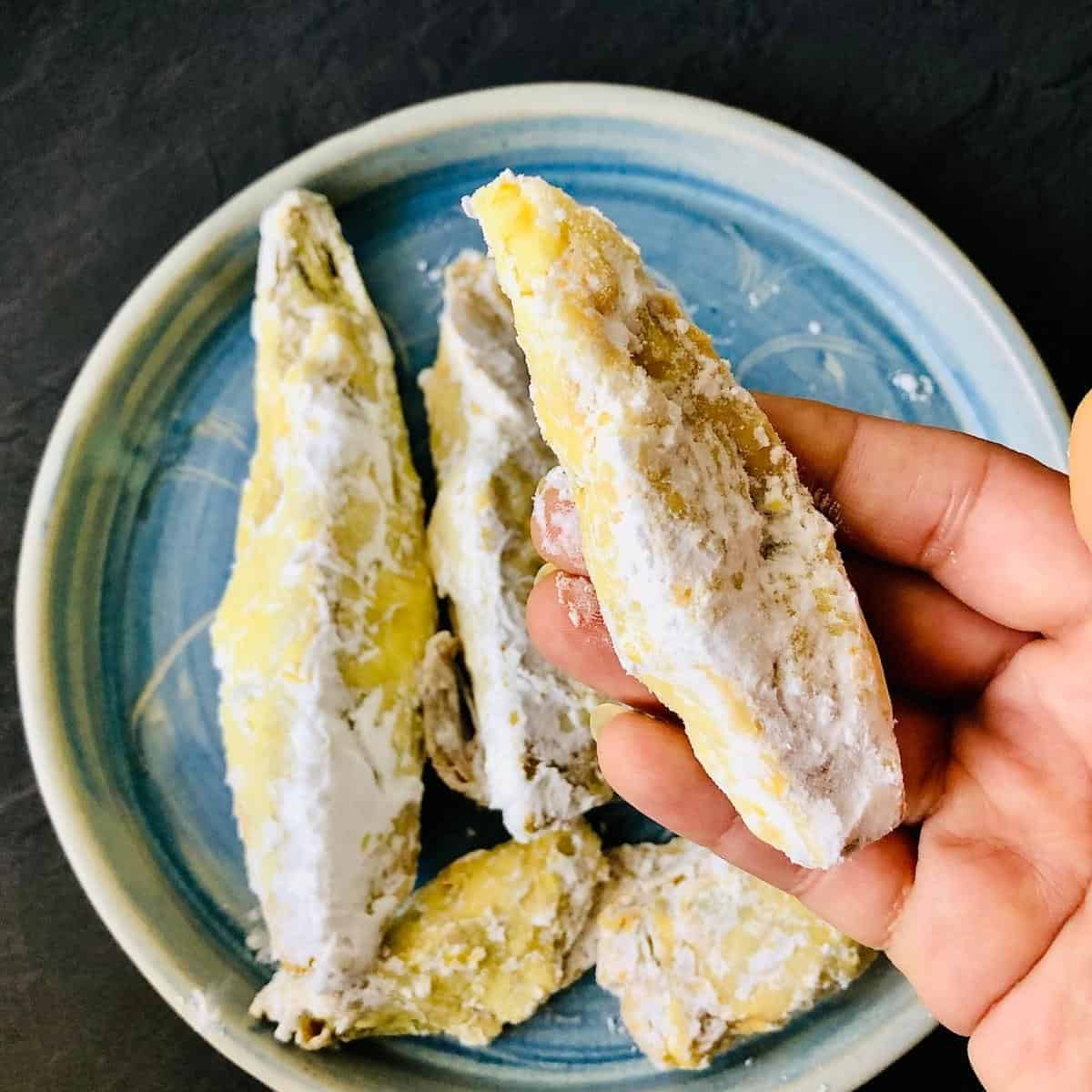 Hand holding a banana blossom coated in potato starch for vegan fish recipe. A plate with other coated banana blossom sits in the background