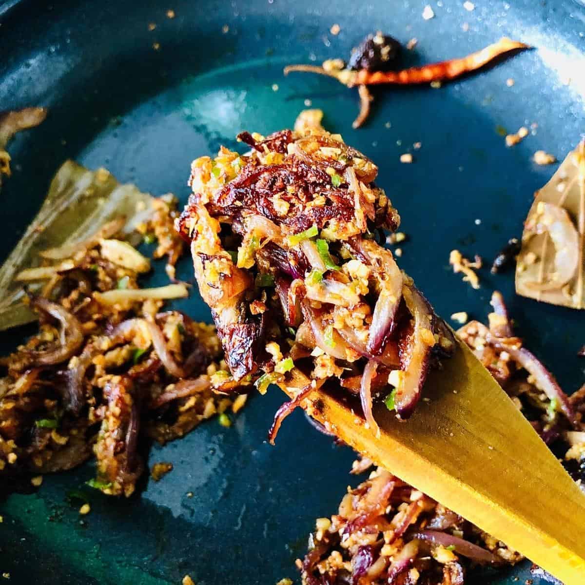 A wooden spoon containing onions, garlic, ginger, green chilli and spices after cooking held over a frying pan
