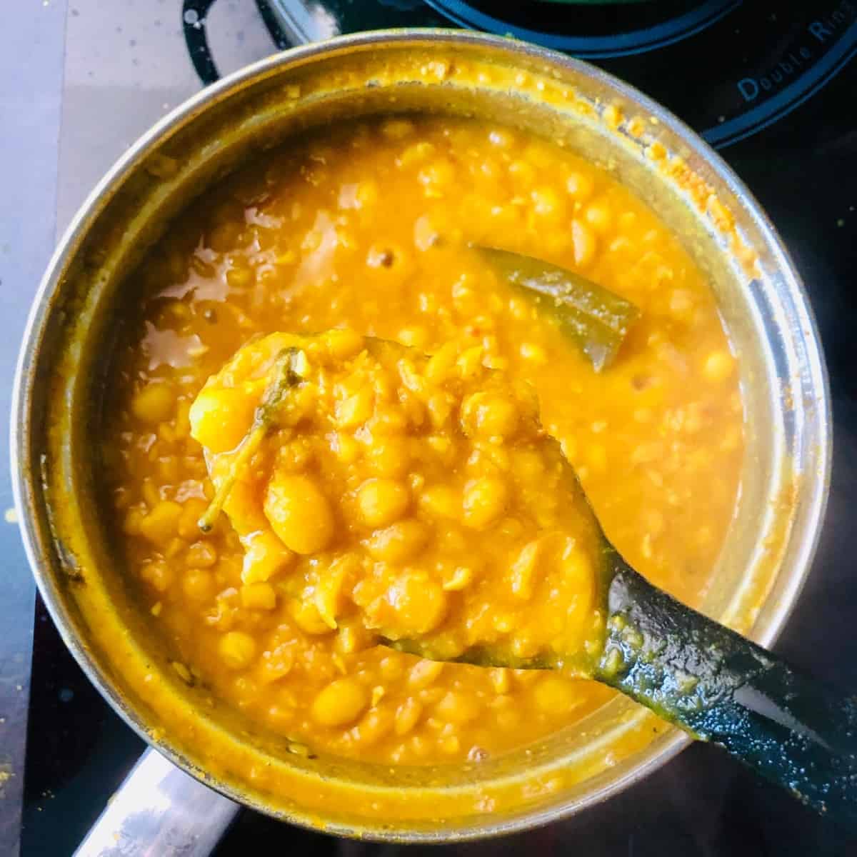 saucepan containing cooked ghugni after half the peas have been crushed to thicken the sauce. A spoon is held above the saucepan containing some of the ghugni