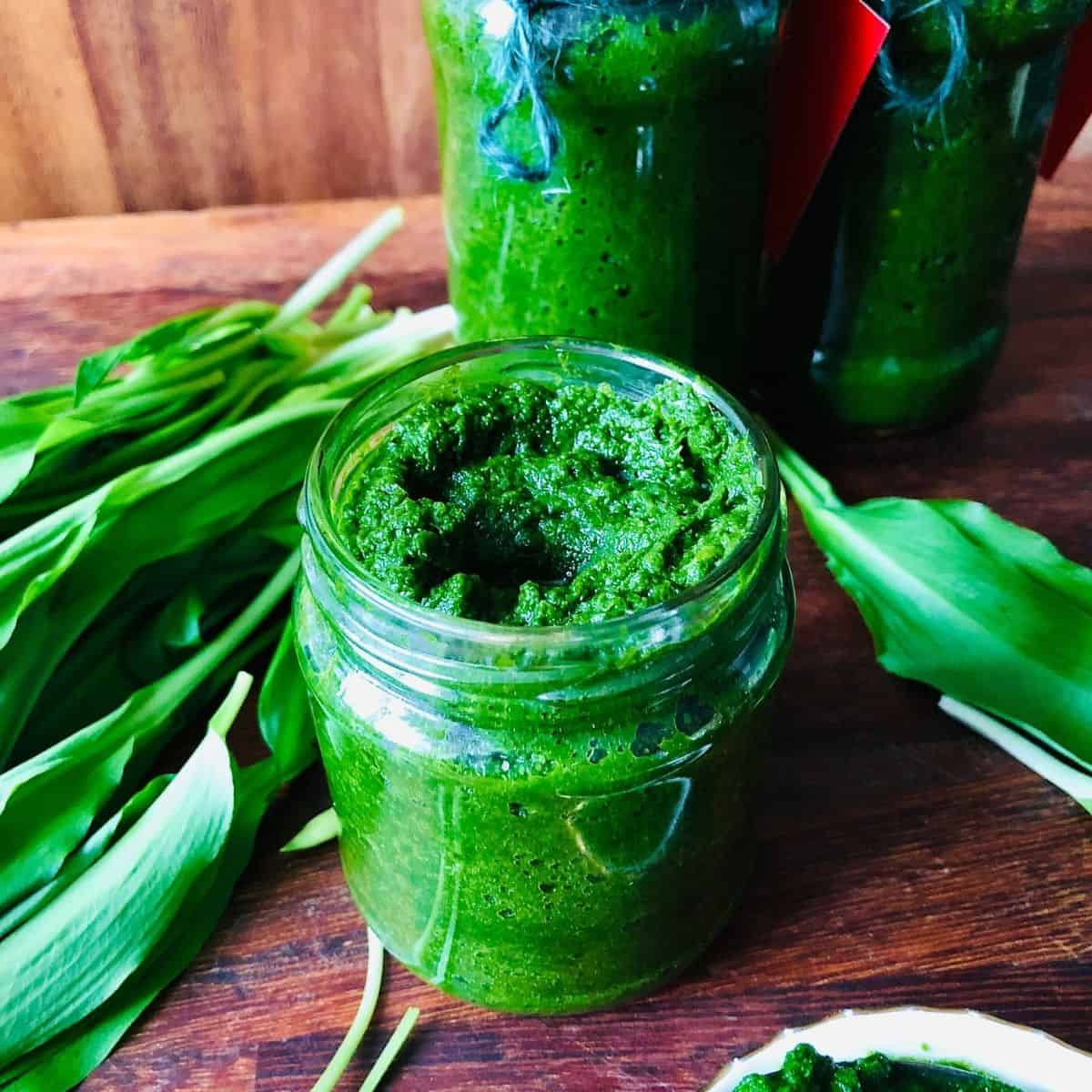 A jar of vivid green wild garlic chilli sauce surrounded by wild garlic leaves