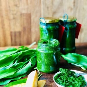 A jar of wild garlic chilli sauce with a small dish of the sauce next to it, surrounded by wild garlic leaves.