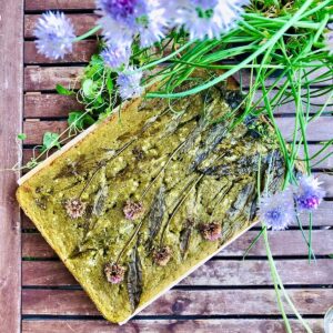 A whole loaf of no knead wild garlic focaccia on a slatted wooden table. Decorated with edible flowers.