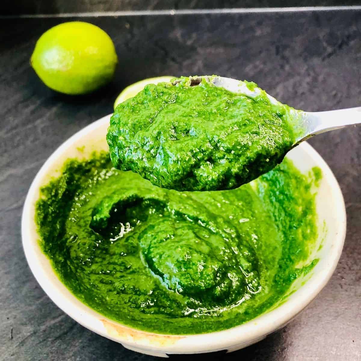 Close up of coriander and chilli chutney in a spoon hovering over a bowl containing the chutney