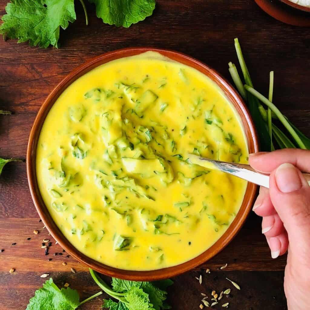 A shallow dish containing kadhi. A hand holds a spoon in the dish