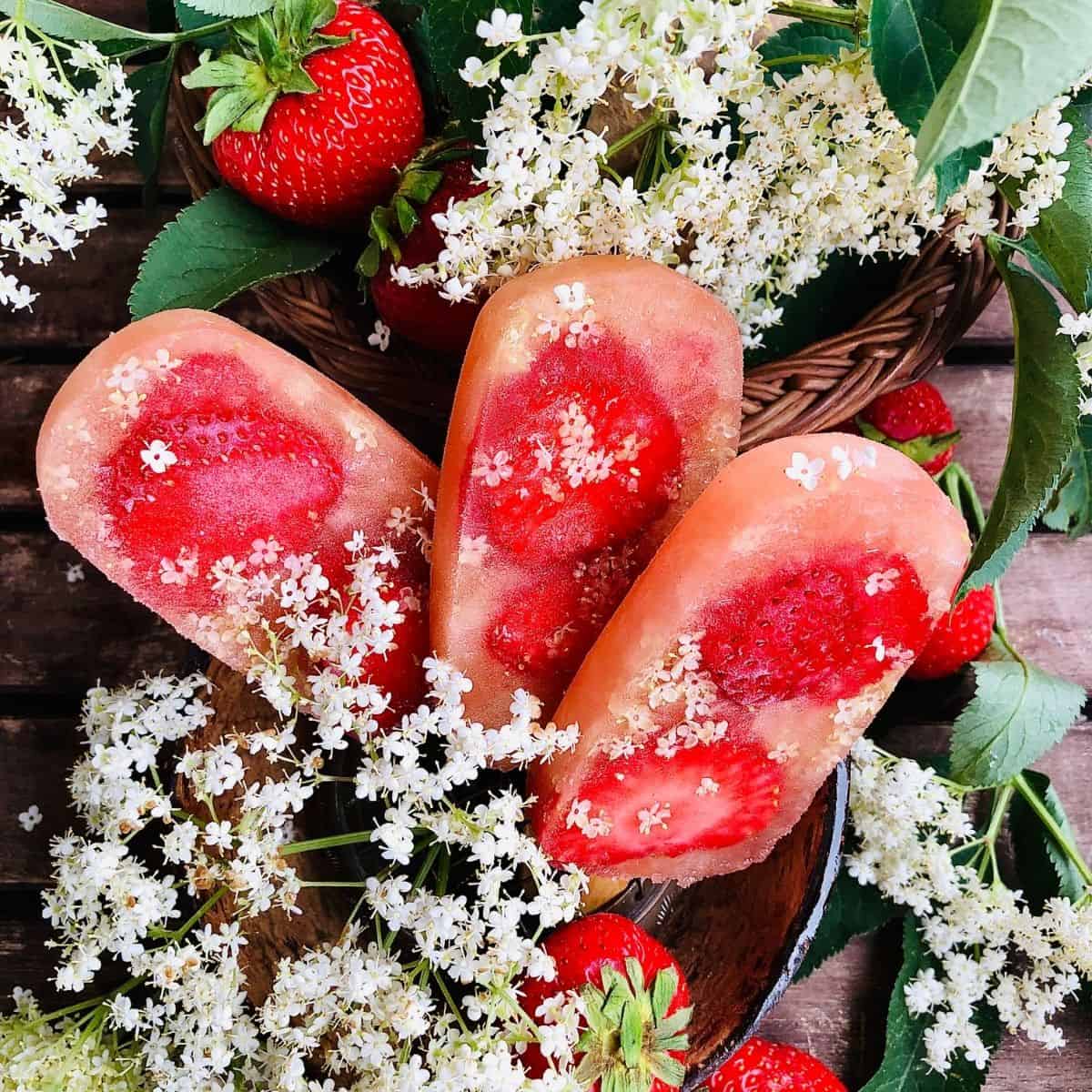 Three elderflower and strawberry ice lollies surrounded by elderflowers and whole strawberries