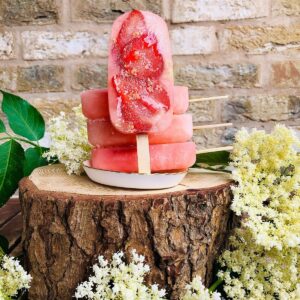 Four Elderflower and strawberry ice lollies stacked on a small dish sat upon a log.