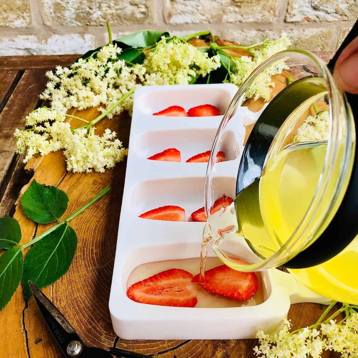 Pouring elderflower cordial into ice lolly mould containing strawberry slices.