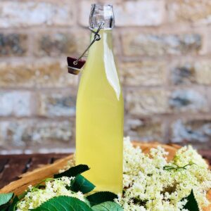 A bottle of elderflower cordial on a wooden platter surrounded by elderflower leaves.