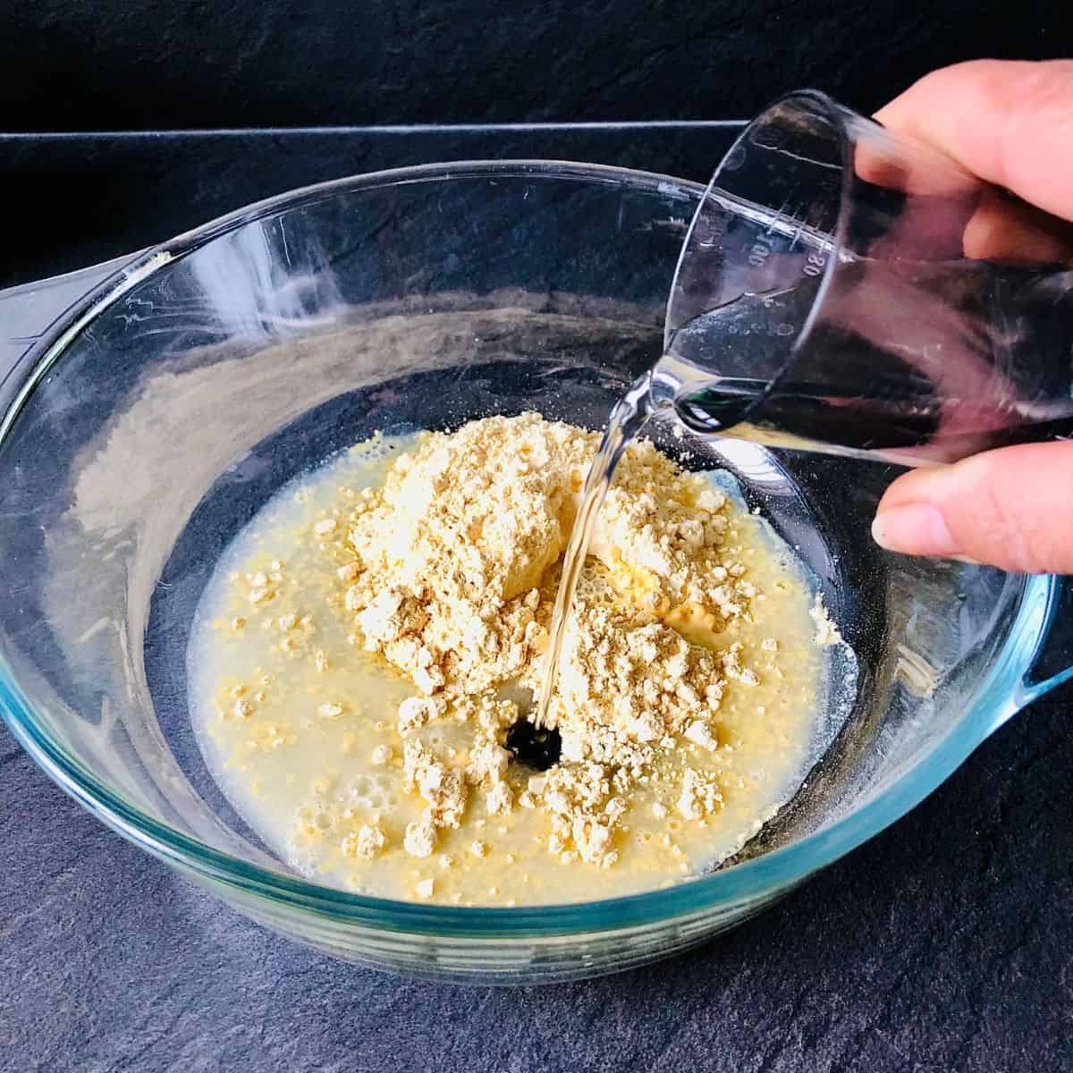 A hand pouring water into a glass bowl containing gram flour to make a batter