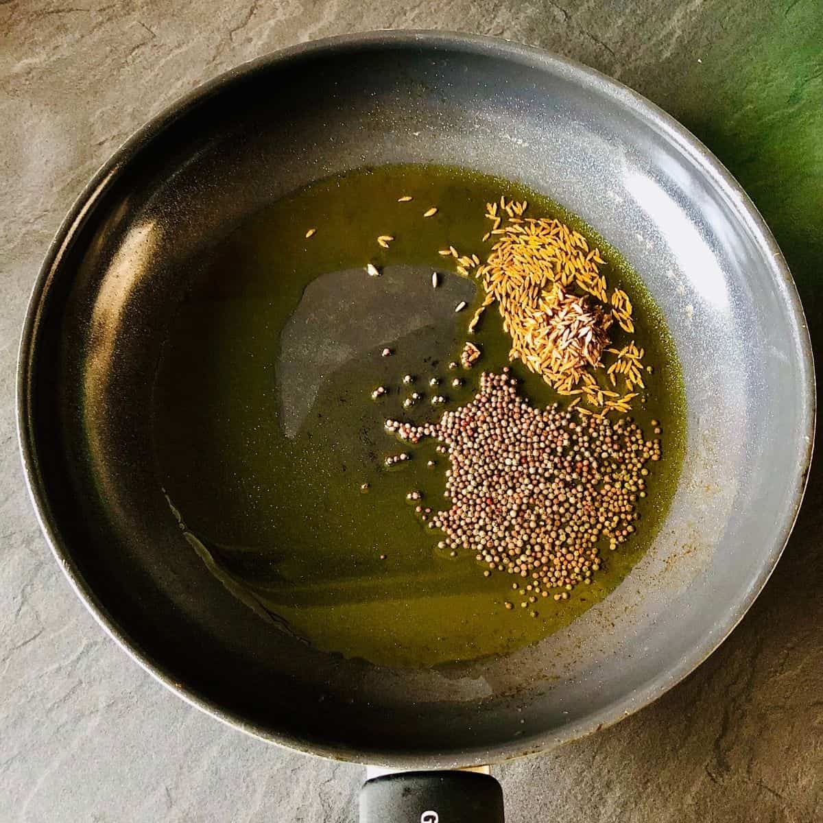 A frying pan containing cumin seeds and mustard seeds frying in mustard oil