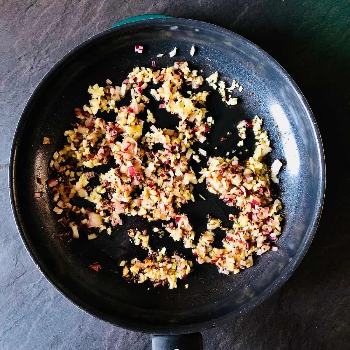 Frying pan containing finely chopped red onion, ginger and garlic, mustard seeds and cumin seeds frying