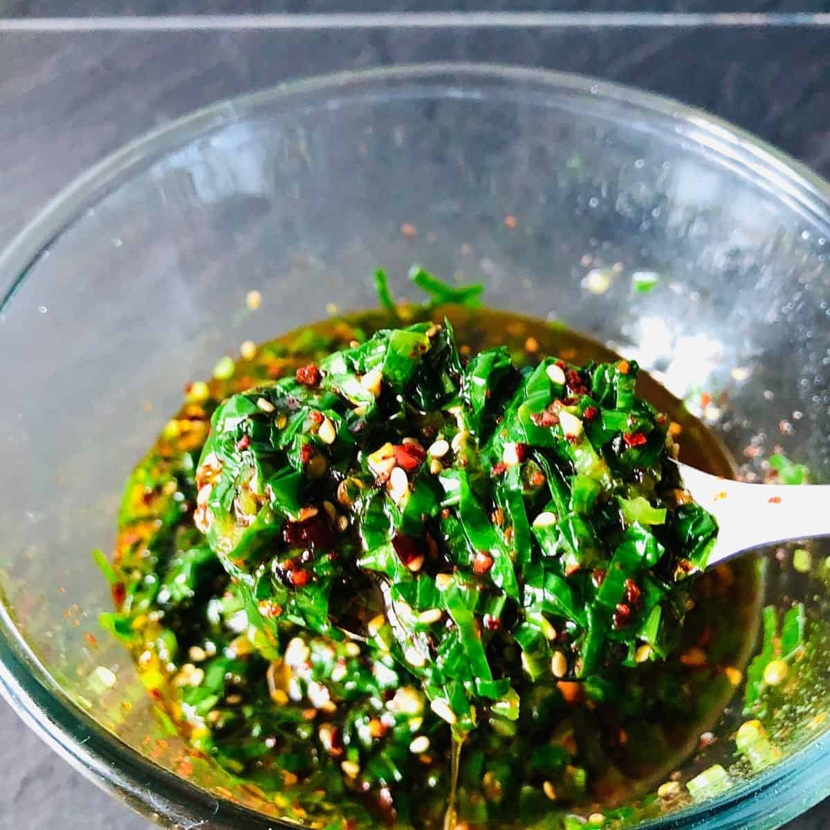 Close up of wild garlic chilli oil on a spoon hovering over a glass bowl containing wild garlic chilli oil