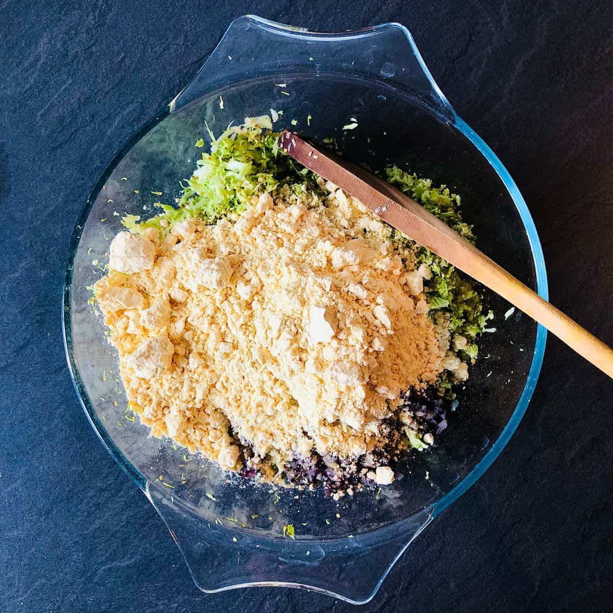 Ingredients for broccoli fritters in a glass bowl ready to mix together with a wooden spoon.