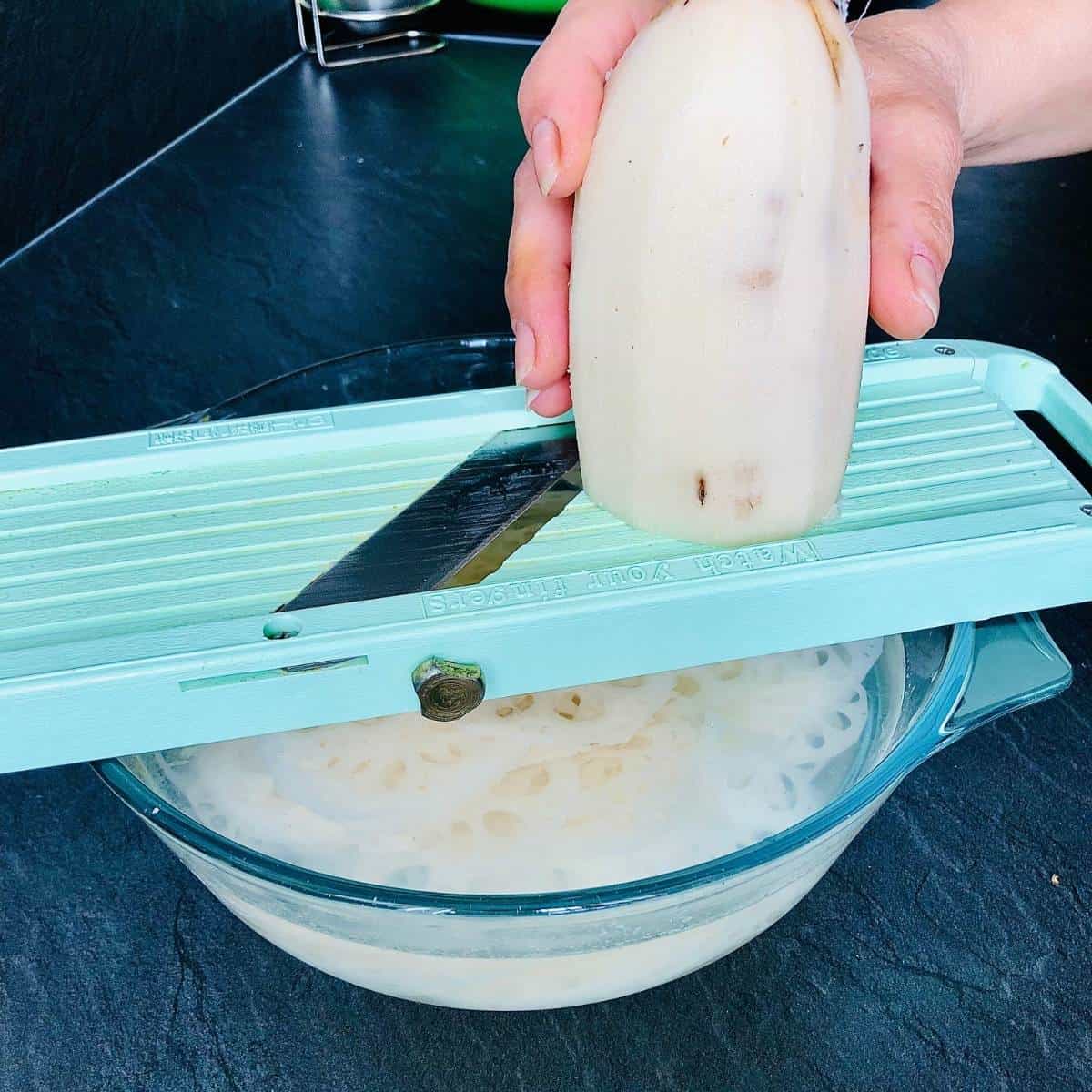 A mandoline slicer resting on a glass dish with a hand passing lotus root over the slicer blade.