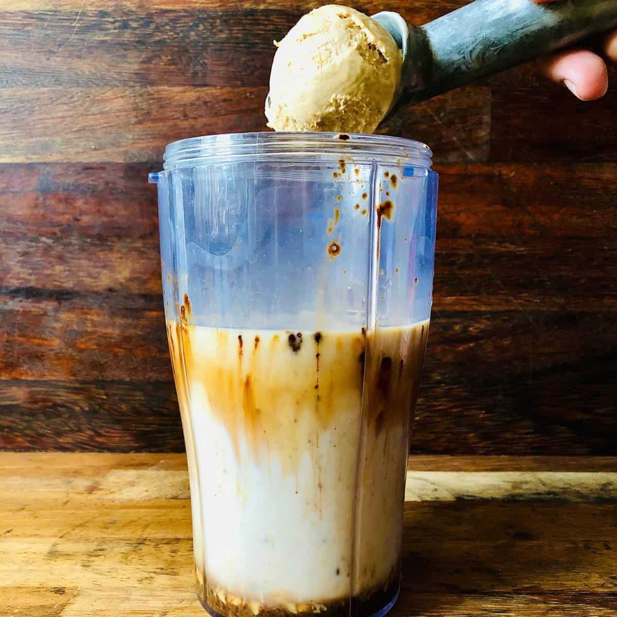 Dropping a scoop of coffee ice cream into a blender cup containing dairy free milk and instant coffee