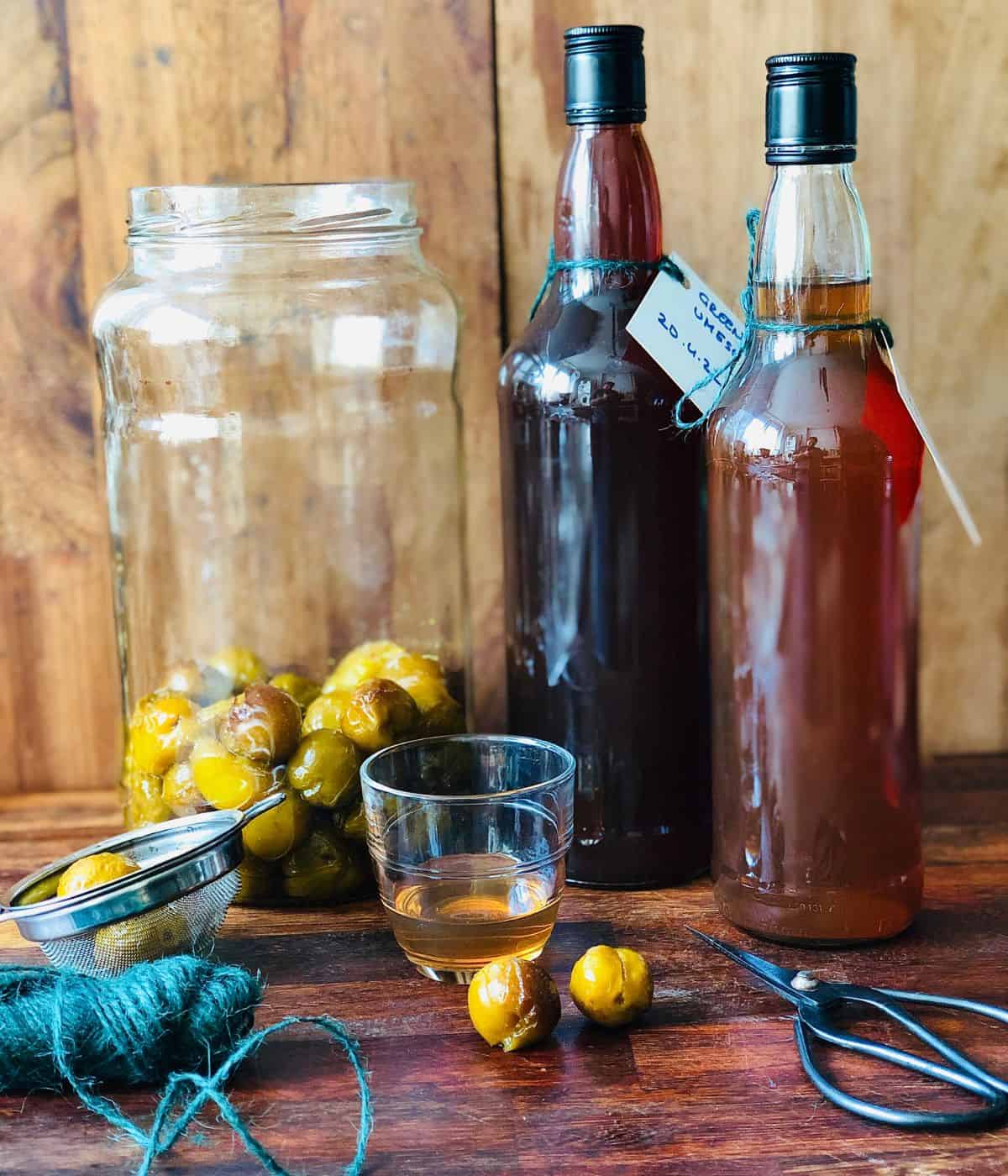 Two bottles of greengage plum wine next to a large jar containing infused fruit and a small glass containing plum wine