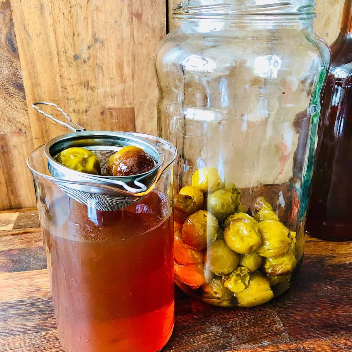 A large jar and a beaker with a strainer on top. The large jar contains alcohol infused greengages. The beaker contains strained plum wine.