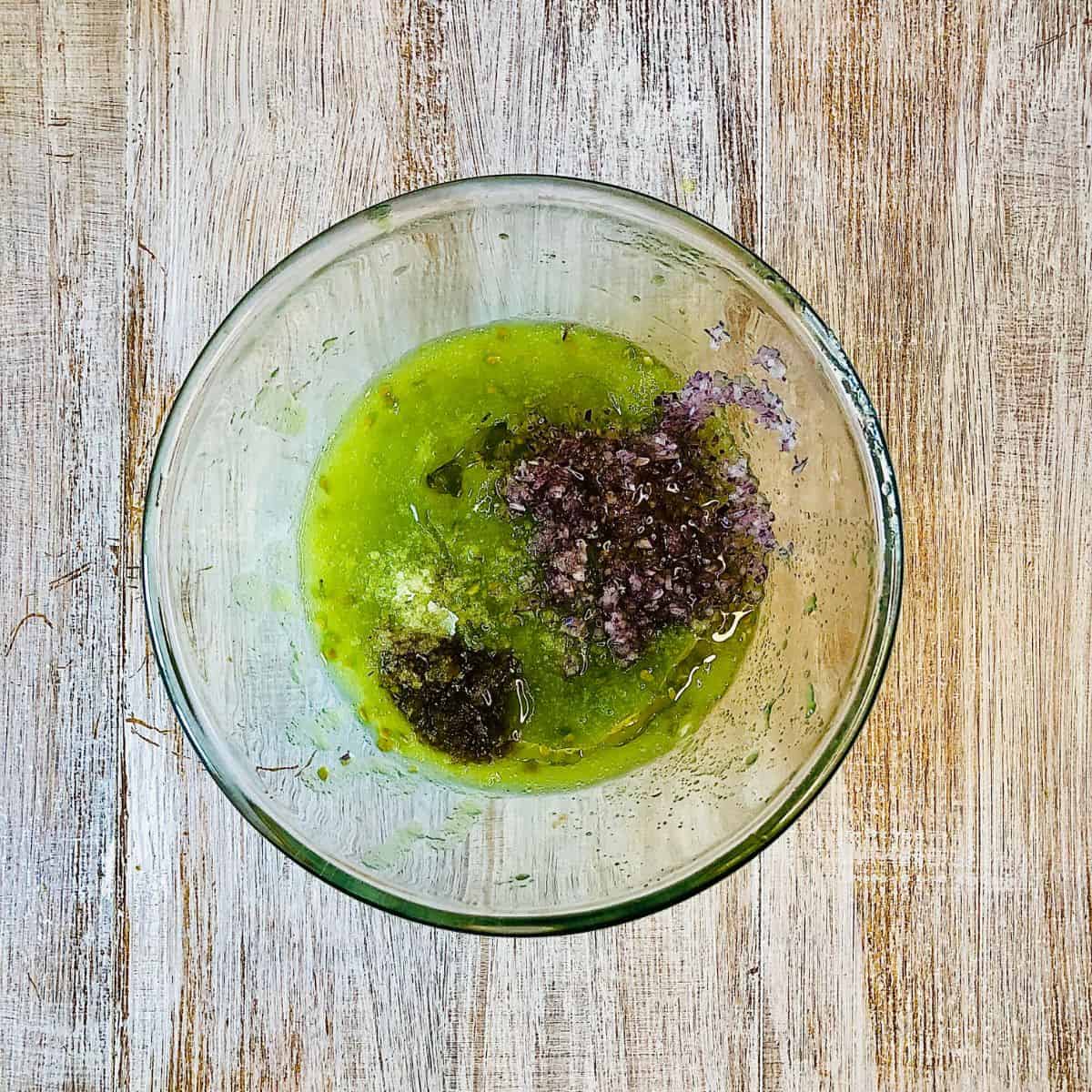 A glass bowl containing grated green heirloom tomato, grated jalapeno pepper, finely chopped red onion and salt, ready to mix.