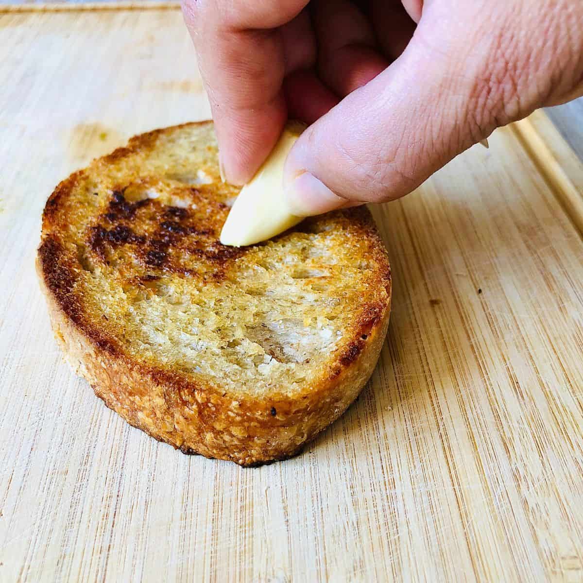 A hand rubbing a piece of garlic over a slice of toasted sourdough bread.