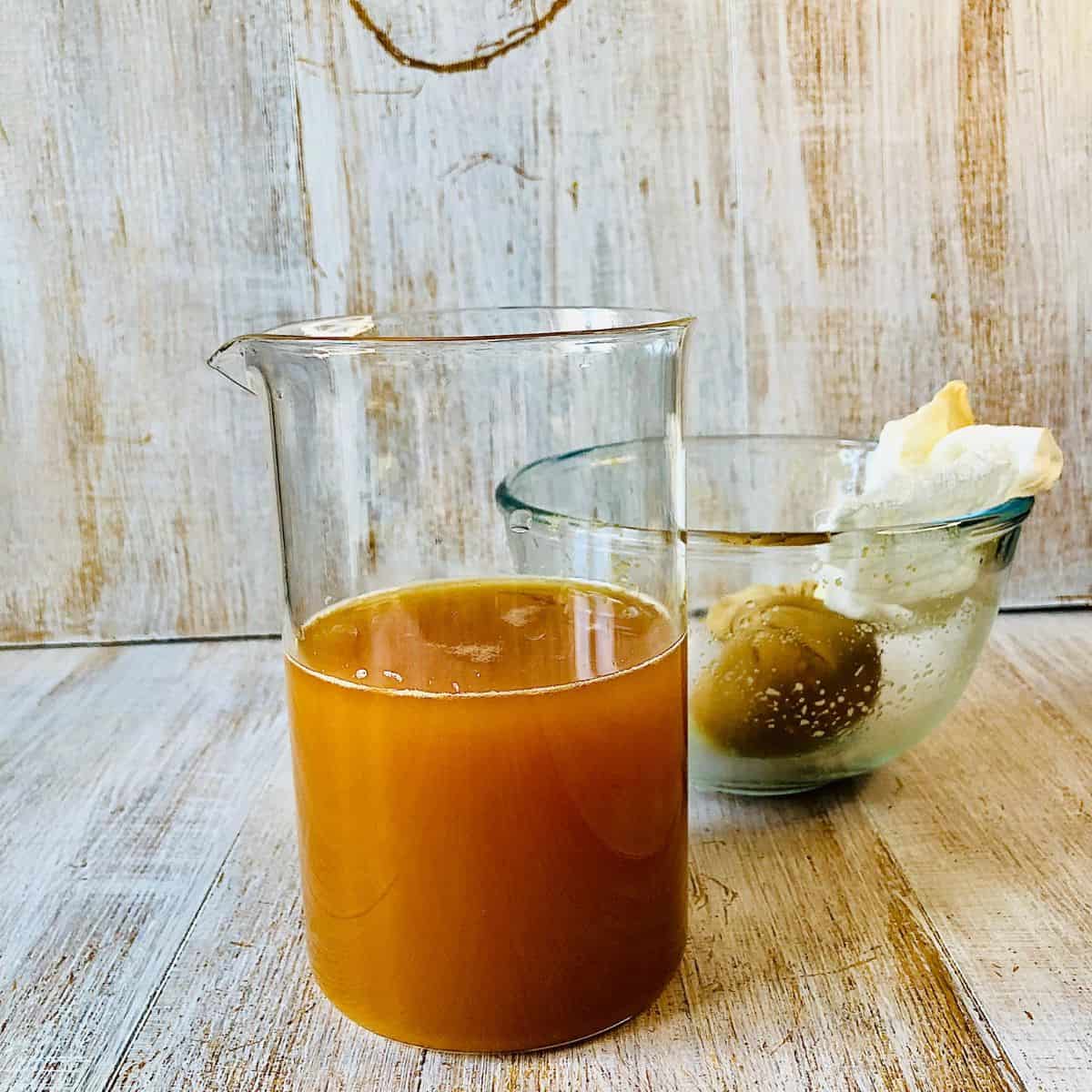 A glass beaker containing apple juice, and a glass bowl containing a muslin cloth with apple pulp in it.