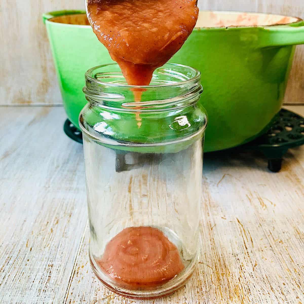 A glass jam jar with crab apple butter being poured into it from a ladle.