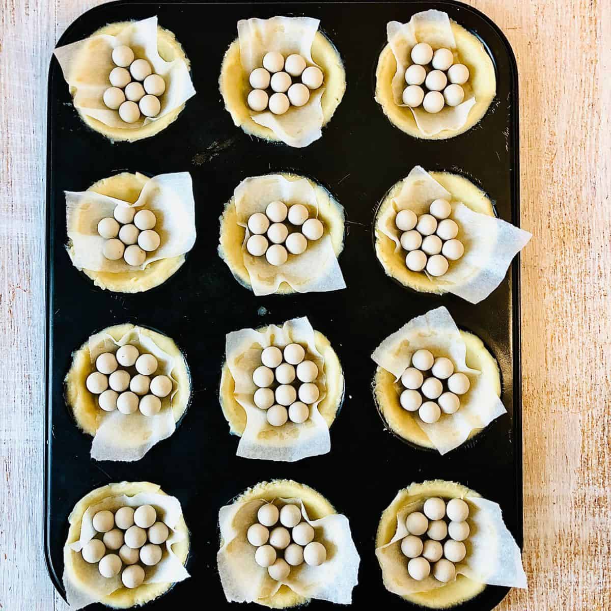 A tray of pastry tart bases with parchment paper and baking balls inside each ready for baking.