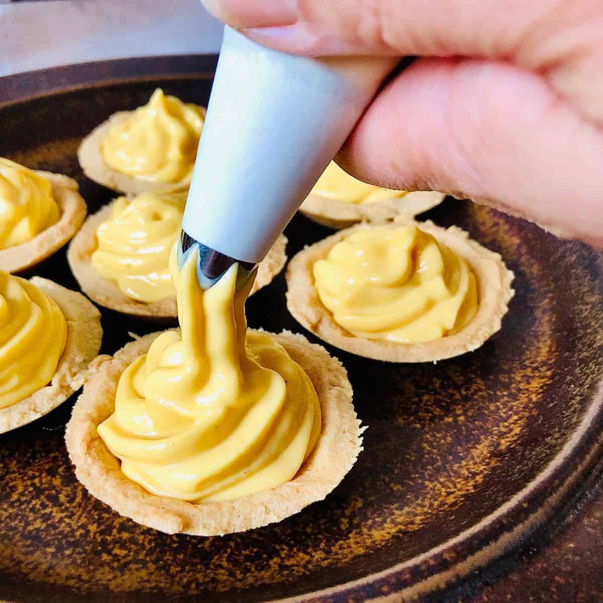 Close up of a piping bag piping mango shrikhand into a pastry base.