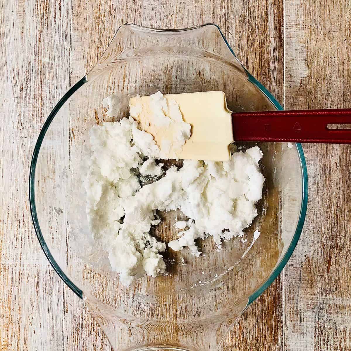 A glass bowl containing a mixture of icing sugar and coconut oil, with a spatula.