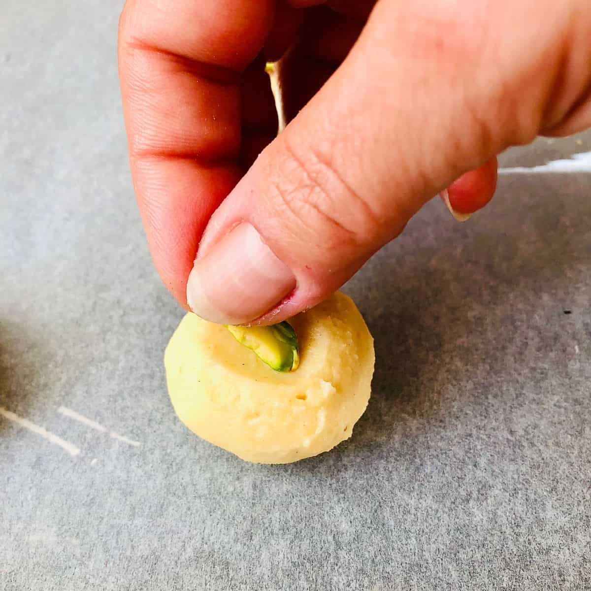 Close up of fingers placing half a pistachio nut into an indentation in the top of a nankhatai biscuit dough ball.