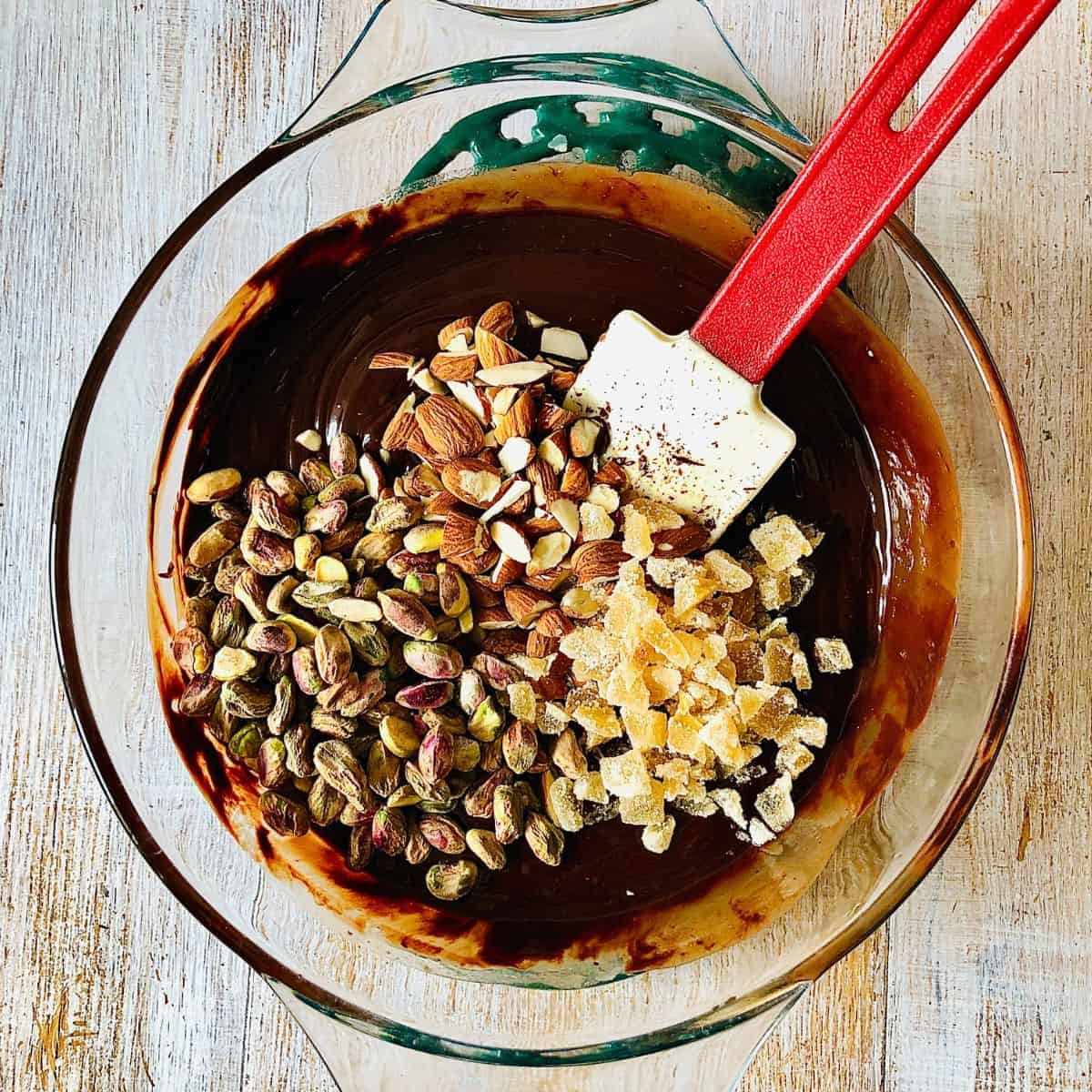 Adding pistachio nuts, almond and crystallised ginger to rocky road mix in a glass bowl.