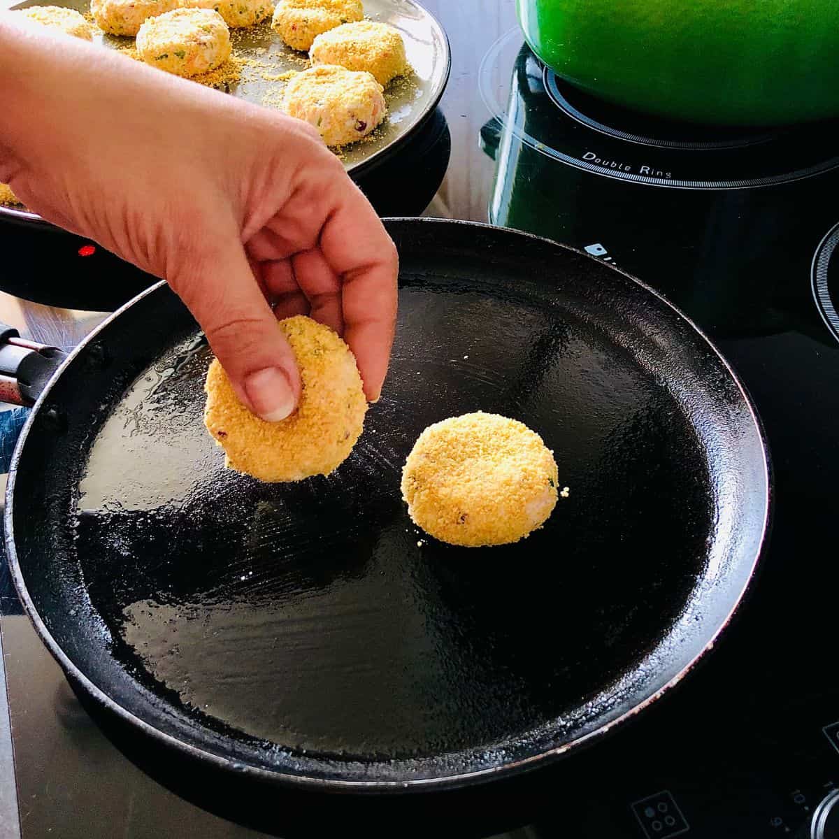 A hand placing a dahi ke kabab patty into a frying pan lightly coated with oil for shallow frying.