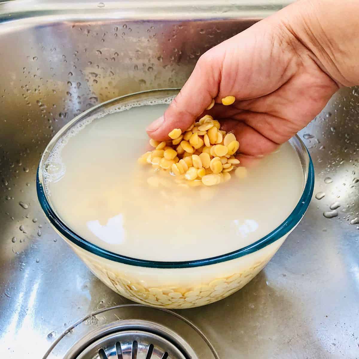 A glass bowl in a sink containing daal in cloudy water before rinsing.