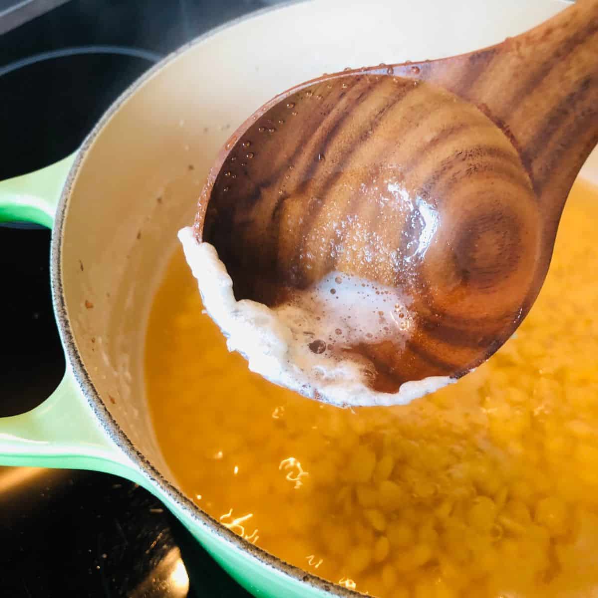 Close up of a wooden spoon containing foam from the surface of daal being cooked. The spoon is held over the pot of cooking daal.